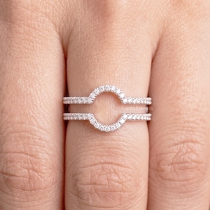 May include: A silver-toned ring guard featuring two parallel bands connected by a central arched bridge. The entire surface of the bands and the arch is encrusted with small, clear, round-cut stones that create a continuous sparkling effect.