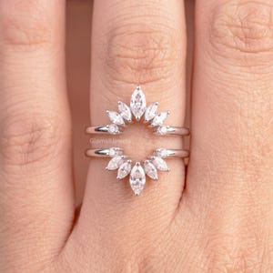 May include: A silver-toned ring guard featuring two curved bands connected by a series of marquise-cut clear stones arranged in a sunburst pattern. The metal is polished and reflective, and the text GlamsiJewels is visible on the center band.