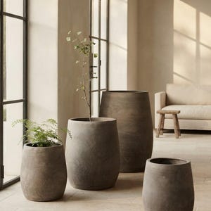 May include: A set of four dark grey, barrel-shaped planters with a smooth, matte finish. Two of the planters contain green plants, while the other two are empty. The planters vary in height and are arranged in a group on a light-coloured floor.