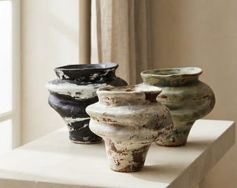 May include: Three ceramic vases with a wide, undulating midsection and a narrow base. One vase is black with white distressed markings, one is off-white with brown textured patches, and one is a muted green with brown and grey weathered details.
