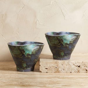 May include: Two ceramic bowls with a flared, irregular rim and a tapered base. The exterior features a dark, textured finish with mottled patches of metallic green and blue glaze, creating a weathered appearance with visible horizontal ridges.