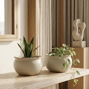 Pode incluir: Dois vasos redondos de cerâmica em tom branco-sujo com detalhes canelados horizontais na metade inferior e uma borda recortada. Um vaso contém uma planta espada-de-são-jorge, o outro uma jiboia verde pendente com longas hastes.