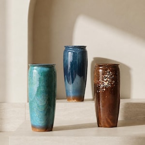 May include: Three cylindrical ceramic vases with a smooth, glossy finish. One vase is turquoise with a brown base, one is deep blue with a lighter blue gradient, and one is brown with a cluster of small, light-colored speckled accents near the top.