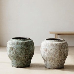 以下が含まれることがあります： 広い口を持つ丸みを帯びた球形の陶器製ジャーが2つ。左側のジャーは青緑色と茶色のまだら模様のテクスチャー仕上げで、右側のジャーは風化したようなオフホワイトとベージュの斑点のある表面の質感が特徴です。