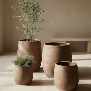 Puede incluir: Un conjunto de cuatro macetas de terracota marrón mate, de formas redondeadas y tamaños variados. Una maceta alta contiene una planta verde esbelta, mientras que una más pequeña alberga una mata de hierba verde. Las macetas tienen lados lisos.