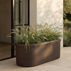Peut inclure: Une grande jardinière de forme ovale avec une finition texturée brun foncé et un rebord arrondi. La jardinière est remplie de hautes herbes ornementales vertes dotées d'épis plumeux de couleur claire dépassant du haut du récipient.