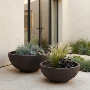 May include: Two large, dark charcoal-coloured, hemispherical planters made of a matte material. The larger planter on the left contains blue-green grasses and succulents, while the smaller planter on the right holds tall, wispy light green grasses and dark purple foliage.