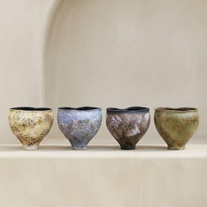 Può includere: Quattro ciotole in ceramica con bordi organici ondulati e basi affusolate. Ogni ciotola presenta una finitura smaltata maculata unica nei toni del crema con macchie marroni, azzurro con riflessi metallici, grigio scuro con toni ruggine e verde oliva.