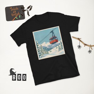 Puede incluir: Camiseta negra con un gr&aacute;fico cuadrado que representa un telef&eacute;rico rojo sobre un fondo de monta&ntilde;as nevadas y cielo azul. La camiseta tiene cuello redondo y mangas cortas. Una peque&ntilde;a pizarra con "Trick or Treat" es visible.