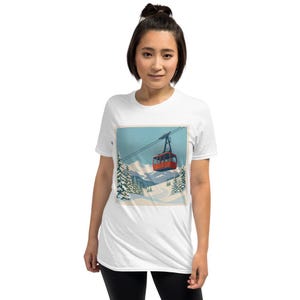 Puede incluir: Camiseta blanca con un gr&aacute;fico de un telef&eacute;rico rojo sobre un paisaje monta&ntilde;oso nevado. El dise&ntilde;o incluye pinos y un cielo azul, evocando un tema de deportes de invierno. La imagen est&aacute; enmarcada.