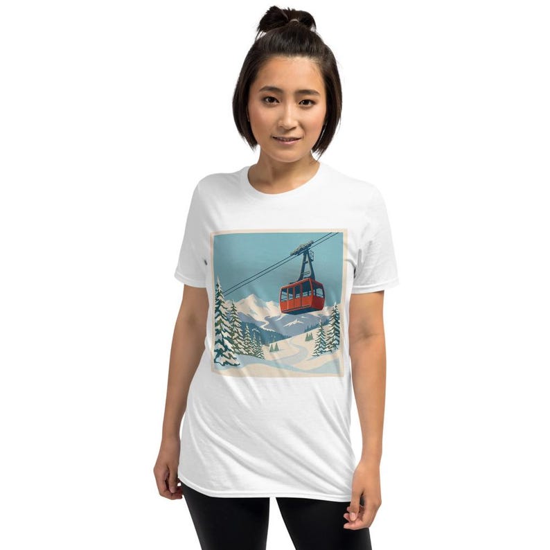 Puede incluir: Camiseta blanca con un gr&aacute;fico de un telef&eacute;rico rojo sobre un paisaje monta&ntilde;oso nevado. El dise&ntilde;o incluye pinos y un cielo azul, evocando un tema de deportes de invierno. La imagen est&aacute; enmarcada.