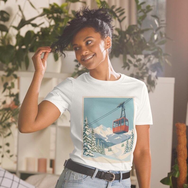Puede incluir: Camiseta blanca con un dise&ntilde;o de telef&eacute;rico vintage. El dise&ntilde;o incluye un telef&eacute;rico rojo, monta&ntilde;as nevadas y pinos bajo un cielo azul. La camiseta la lleva una persona.