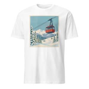 Puede incluir: Camiseta blanca con un estampado retro de un telef&eacute;rico rojo ascendiendo una escena de monta&ntilde;a nevada. El dise&ntilde;o incluye pinos, una pista de esqu&iacute; sinuosa y un cielo azul, que recuerda a un cartel de viaje vintage.