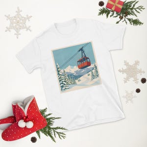 Puede incluir: Camiseta blanca con un gr&aacute;fico de paisaje invernal. El dise&ntilde;o muestra un telef&eacute;rico rojo ascendiendo una monta&ntilde;a nevada, con pinos y cielo azul. La camiseta est&aacute; rodeada de adornos navide&ntilde;os.