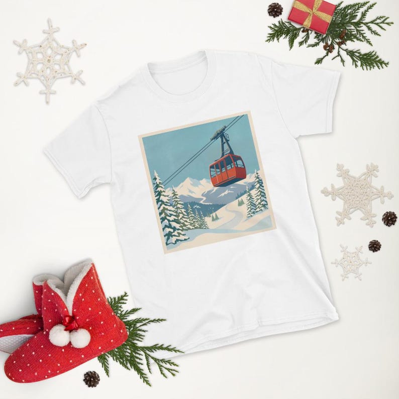 Puede incluir: Camiseta blanca con un gr&aacute;fico de paisaje invernal. El dise&ntilde;o muestra un telef&eacute;rico rojo ascendiendo una monta&ntilde;a nevada, con pinos y cielo azul. La camiseta est&aacute; rodeada de adornos navide&ntilde;os.