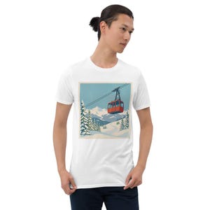 Puede incluir: Camiseta blanca con una ilustraci&oacute;n de estilo retro de un telef&eacute;rico rojo ascendiendo una monta&ntilde;a nevada. El dise&ntilde;o incluye pinos cubiertos de nieve y un cielo azul, evocando un tema de deportes de invierno.