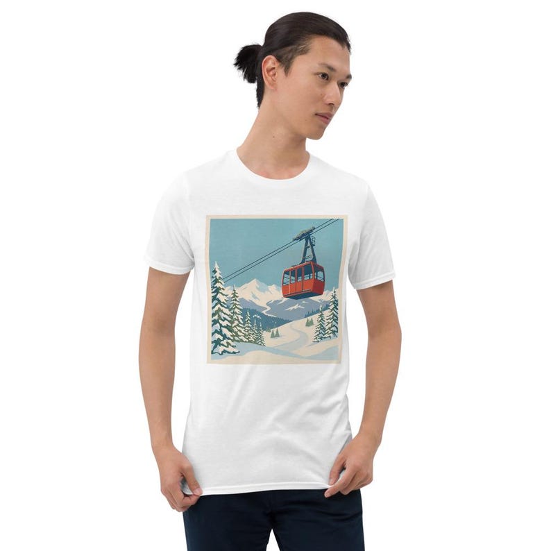 Puede incluir: Camiseta blanca con una ilustraci&oacute;n de estilo retro de un telef&eacute;rico rojo ascendiendo una monta&ntilde;a nevada. El dise&ntilde;o incluye pinos cubiertos de nieve y un cielo azul, evocando un tema de deportes de invierno.