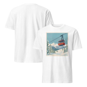 Puede incluir: Camiseta blanca con un estampado gr&aacute;fico de un telef&eacute;rico rojo sobre un fondo de monta&ntilde;a nevada. El dise&ntilde;o incluye pinos y un cielo azul, evocando una escena invernal. La camiseta est&aacute; hecha de un material suave.