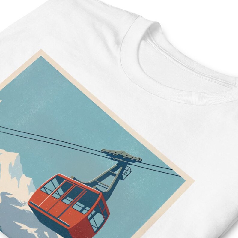 Puede incluir: Camiseta blanca con una ilustraci&oacute;n de estilo retro de un telef&eacute;rico rojo sobre un fondo de cielo azul y monta&ntilde;as nevadas. El dise&ntilde;o tiene una est&eacute;tica de cartel de viaje vintage, que evoca aventura y aire libre.