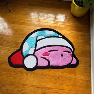 May include: A pink, black, and white rug shaped like a cartoon character wearing a blue and white polka-dot hat. The rug has a red section and is lying on a wooden floor.
