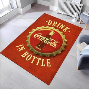 May include: A rectangular area rug featuring a vibrant red background with a large, central gold-toned bottle cap graphic. The design includes a classic Coca-Cola glass bottle illustration and the text DRINK COCA-COLA IN BOTTLE in bold lettering.