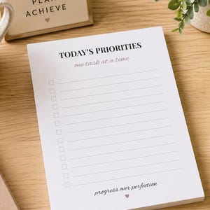 May include: A notepad with the text "TODAY'S PRIORITIES" and "one task at a time" is on a wooden desk. A mug of coffee, a keyboard, a pen, and a notebook with glasses are also on the desk. The notepad also has the text "progress over perfection".