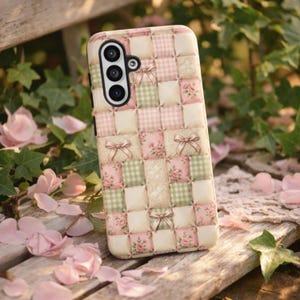 Puede incluir: Una funda para smartphone con un diseño de patchwork acolchado que alterna cuadrados en color crema, cuadros vichy verde salvia y estampados florales rosas, decorada con pequeños lazos beige. La funda tiene un módulo de cámara negro con tres lentes.