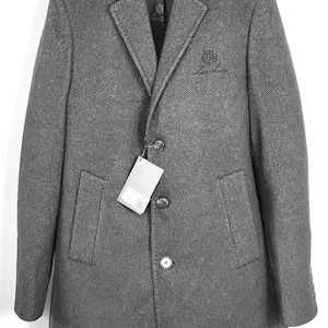 May include: A charcoal gray overcoat featuring a subtle diagonal weave pattern, a notched lapel, and a three-button front closure. The coat includes two side pockets, a small embroidered logo on the chest, and a brand tag attached to the middle button.