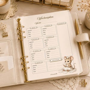 Könnte beinhalten: Ein beigefarbener Ringplaner mit einer Wochenplan-Einlage. Die Seite enthält Felder für Montag bis Sonntag, eine Einkaufsliste und eine kleine Maus-Illustration. Ein goldener Stift ist in den Einband des Planers eingesteckt.