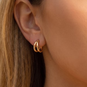 May include: A pair of small, polished gold hoop earrings featuring a double-layered design. The earrings are circular in shape with a smooth, reflective metallic finish, creating a stacked appearance when worn in the earlobe.