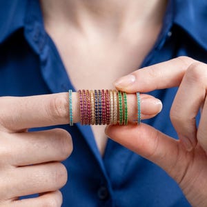 May include: A collection of thin, stackable metal rings featuring a continuous row of small, round-cut gemstones in various colors including turquoise, pink, red, dark blue, clear white, and green, all set in a polished gold-tone metal band.