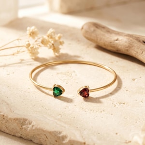 Può includere: Un bracciale aperto color oro con due pietre preziose a forma di cuore alle estremità. Una pietra è di un vibrante verde smeraldo, mentre l'altra è di un profondo rosso granato. Il sottile cinturino in metallo lucido crea un design simmetrico.