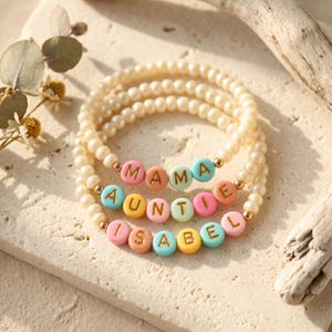 Könnte beinhalten: Drei elastische Armbänder aus kleinen weißen Kunstperlen. Jedes Armband enthält bunte runde Perlen mit goldenen Buchstaben, die MAMA, AUNTIE und ISABEL buchstabieren, unterbrochen von kleinen goldfarbenen Metallzwischenperlen.