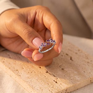 Può includere: Un anello aperto color argento con un gruppo di pietre ametista viola taglio marquise e piccole pietre trasparenti disposte a forma di foglia sulla parte superiore della fascia, creando un gioiello delicato di ispirazione botanica.