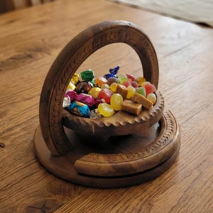 Op de afbeelding: Een houten snoepmand gevuld met kleurrijk verpakt snoep en onverpakt snoep. De mand heeft een gesneden handvat en een decoratieve basis, rustend op een houten oppervlak. De snoepjes omvatten karamels, gelei snoepjes en chocolaatjes.