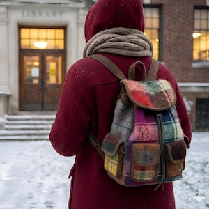 May include: A multi-colored plaid backpack with three front pockets and a drawstring closure, worn over a deep red hooded coat with a beige knit scarf. The backpack features a mix of red, green, blue, and yellow wool-like fabric patterns.