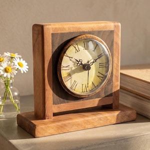 May include: A rectangular wooden table clock with a light brown frame and a dark brown centre panel. The round clock face features a cream-coloured background, black Roman numerals, and black ornate hands protected by a clear glass cover.