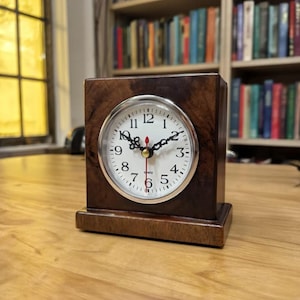 May include: A square wooden desk clock with a white clock face and black hands. The clock is set in a dark brown wooden frame and sits on a wooden base. The clock face has black numbers and a red second hand.