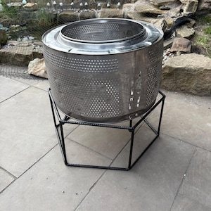 May include: A silver metal fire pit, crafted from a repurposed washing machine drum, mounted on a black metal frame. The drum has many small holes. The fire pit is set on a stone patio, with a pond and rocks visible in the background.