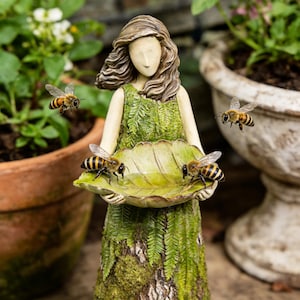 Pode incluir: Estátua de jardim de uma mulher com um vestido verde segurando um bebedouro em forma de folha. Várias abelhas voam ao redor da estátua e bebem água. A estátua tem uma aparência desgastada e natural, ideal para decoração ao ar livre.
