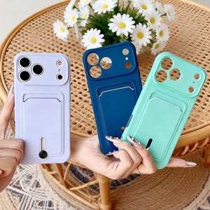 May include: Three mobile phone cases in light purple, dark blue, and mint green. Each case features a rectangular card slot on the back and a circular cutout for the camera lens array. The purple case includes a small black sliding button feature.