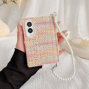 Op de afbeelding: Een rechthoekig telefoonhoesje met een geweven tweed-achtige textuur in pasteltinten van roze, geel en blauw. Het hoesje is voorzien van een wit parelarmbandje bevestigd aan een zilverkleurige metalen sluiting en een witte camerabehuizing.