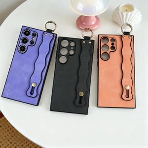May include: Three phone cases in lavender, black, and peach. Each case has a wavy strap, a gold-tone ring, and a gold-tone button. The cases have cutouts for the phone's camera and buttons.
