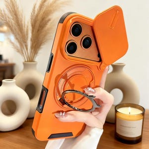 Op de afbeelding: Oranje telefoonhoesje met een cameraklepje en een zilveren ringhouder. De hoes heeft zwarte accenten en een verhoogde rand. De ringhouder zorgt voor een stevige grip. De hoes is ontworpen om de camera van de telefoon te beschermen.