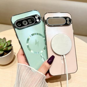Può includere: Due custodie per telefono, una verde menta e una rosa, sono tenute in mano. La custodia verde menta ha un design a fulmine argentato e un ritaglio per la fotocamera. La custodia rosa ha un pad di ricarica circolare bianco e un cavo di ricarica.
