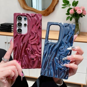 May include: Two glossy phone cases, one in a deep magenta and the other in a vibrant blue. Both cases have a textured, rippled surface. The magenta case has multiple camera lenses, while the blue case has a single camera opening.