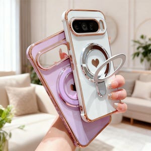 May include: Two phone cases are displayed, one white and one lavender. The white case has a gold-coloured frame and a silver ring with a heart-shaped cutout. The lavender case has a gold-coloured frame and a silver ring with a heart-shaped cutout.