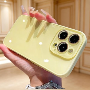Puede incluir: Una funda de teléfono amarillo pálido con un acabado brillante. La funda tiene un módulo de cámara cuadrado con tres lentes negras y un borde dorado. La funda se sostiene en una mano, mostrando su diseño y color.