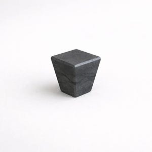 Puede incluir: Un tirador de armario geométrico gris oscuro, con una parte superior cuadrada y un cuerpo trapezoidal. El tirador está hecho de un material sólido y texturizado y está sobre un fondo blanco. El diseño es moderno y minimalista.