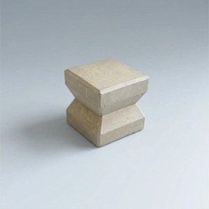Op de afbeelding: Een lichtbeige, geometrische betonnen kruk met een unieke zandloper vorm. De kruk heeft een vierkante boven- en onderkant, verbonden door een smaller, schuin middengedeelte. De neutrale kleur en het moderne ontwerp maken het een veelzijdig stuk.